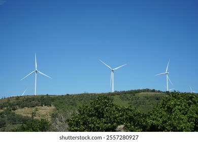 This image shows a landscape with wind turbines on a hill, surrounded by green trees, under a clear blue sky. This location is in Sidrap Regency, South Sulawesi.