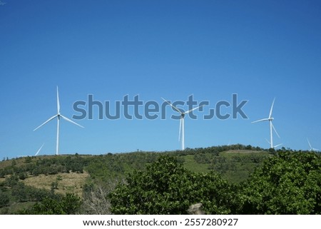 This image shows a landscape with wind turbines on a hill, surrounded by green trees, under a clear blue sky. This location is in Sidrap Regency, South Sulawesi.