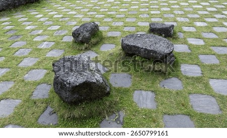 This image shows large, dark, weathered rocks scattered on a grassy area, interspersed with a grid of square stepping stones, creating an informal garden or pathway