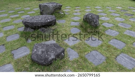 This image shows large, dark, weathered rocks scattered on a grassy area, interspersed with a grid of square stepping stones, creating an informal garden or pathway