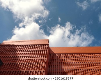 This image shows a low angle view looking up at a modern building facade featuring large decorative ventilation screens made of concrete roaster blocks. The entire exterior structure, including the solid wall panels and the hollow ventilation bricks, is painted in a uniform terracotta orange color. The facade contains massive grids of repeating vertical rectangular openings designed for airflow and shade. Bright daylight casts strong geometric shadows across parts of the structure, highlighting its architectural corners and straight lines against a vibrant blue sky filled with scattered white fluffy clouds, presenting a contemporary tropical architectural design.