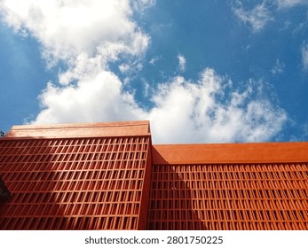 This image shows a low angle view looking up at a modern building facade featuring large decorative ventilation screens made of concrete roaster blocks. The entire exterior structure, including the solid wall panels and the hollow ventilation bricks, is painted in a uniform terracotta orange color. The facade contains massive grids of repeating vertical rectangular openings designed for airflow and shade. Bright daylight casts strong geometric shadows across parts of the structure, highlighting its architectural corners and straight lines against a vibrant blue sky filled with scattered white fluffy clouds, presenting a contemporary tropical architectural design.