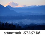 This image shows a panoramic view of layered mountains covered in a light mist with calming blue nuances. Silhouettes of hills and mountains appear to rise into the distance, creating an impression of breadth and depth. Below you can see rows of dark trees faintly covered in morning dew, while soft clouds hang over the mountains with a touch of pale orange from the sunlight. The atmosphere in the picture feels cool, quiet and peaceful, depicting the natural beauty of the mountains at dawn or near dusk.