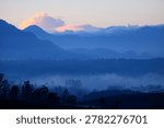 This image shows a panoramic view of layered mountains covered in a light mist with calming blue nuances. Silhouettes of hills and mountains appear to rise into the distance, creating an impression of breadth and depth. Below you can see rows of dark trees faintly covered in morning dew, while soft clouds hang over the mountains with a touch of pale orange from the sunlight. The atmosphere in the picture feels cool, quiet and peaceful, depicting the natural beauty of the mountains at dawn or near dusk.
