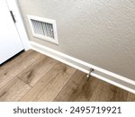 This image shows a section of a room with new wood flooring. The focus is on a white floor vent and a door stop installed on the baseboard. The vent has horizontal slats and is positioned close to the