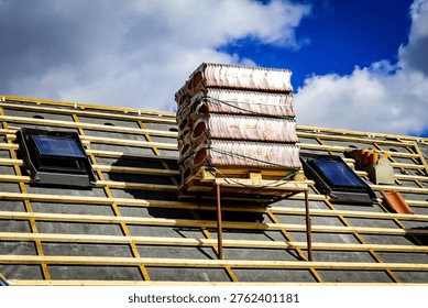 This image shows a small section of a renovated roof on a five-story residential apartment building. Skylight windows have been installed, and roof tiles are stacked on a pallet placed on a metal support structure on the roof, ready to be installed to complete the roof renovation. The image documents the final stage of a roofing project on a multi-story residential building, focusing on roofing materials, construction details, and renovation work in progress.
