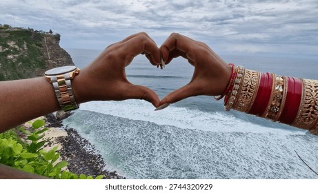 This image shows two hands forming a heart shape in the foreground, framing a beautiful ocean view in the background, The hands are joined together to create a heart symbol.