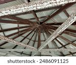 This image shows the underside of a circular or radial roof structure. The frame is made of metal beams that converge at a central point, creating a star-like pattern. The beams are heavily rusted and