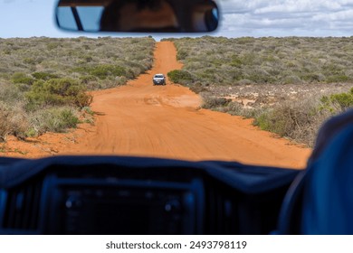 This image shows a view from inside a vehicle on a dirt road surrounded by green shrubs and an orange sandy path leading into the distance under a partly cloudy sky.