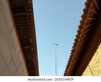 This image shows a view looking up between two buildings with tiled and corrugated roofs. In the center, a tall pole with a TV antenna is silhouetted against a clear blue sky. The sunlight casts sharp