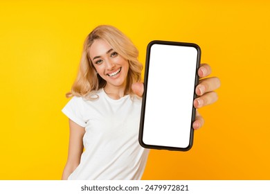 This image shows a woman with blonde hair smiling while holding up a smartphone with a blank screen. The woman is wearing a white t-shirt and is standing in front of a bright yellow background. - Powered by Shutterstock