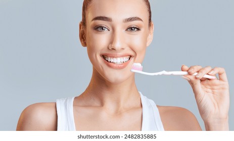 This image shows a woman with a white tank top brushing her teeth with a toothbrush and toothpaste. She has a bright smile and appears happy and healthy.