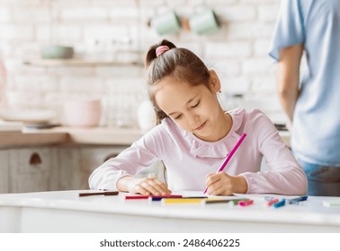 This image shows a young girl sitting at a white table in her home, focusing intently on a drawing she is coloring with a pink crayon. There are other crayons and markers scattered on the table - Powered by Shutterstock