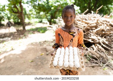 In This Image, A Smart Black Girl With Big Eyes And Gentle Smile, Is Standing In Font Of Bundles Of Firewood, With A Tray Full Of Eggs