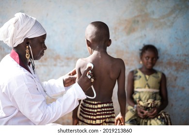 In This Image A Smiling Black Doctor Sitting In A Classroom Is Auscultating One Of Two Little African Children With A Stethoscope During COVID School Screening