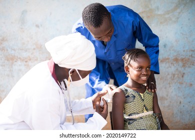 In this image, a smiling cute African girl is being injected a vaccine dose in her upper arm by a black female pediatrician with mask and white uniform assisted by a blue coated healthcare worker - Powered by Shutterstock