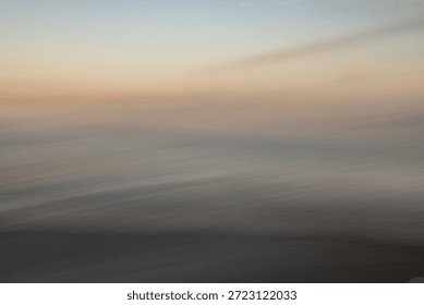 This image is a sophisticated example of Intentional Camera Movement (ICM) photography, capturing a serene and ethereal seascape at the transition of twilight. The composition is defined by its soft, painterly quality, where the traditional boundaries between sea, sky, and shore are dissolved into a fluid arrangement of horizontal bands. The color palette is exceptionally delicate, featuring a gradient of muted oceanic blues, hazy grays, and a warm, glowing amber horizon that suggests the final moments of a sunset or the first light of dawn.

The long exposure and deliberate panning motion create a rhythmic sense of movement, transforming the crashing waves into a series of soft, atmospheric streaks. This abstraction emphasizes the emotional resonance of the coast—tranquility, solitude, and the ephemeral nature of time—rather than its literal details. The subtle textures within the blur give the piece a tactile, canvas-like feel, making it an ideal choice for high-end interior decor, meditative editorial content, or minimalist graphic design backgrounds.

There is a profound sense of stillness despite the implied motion. The heavy, dark tones at the bottom provide a grounded foundation, while the lighter, airy hues at the top evoke a sense of vastness and possibility. This balance of light and shadow, combined with the dreamlike aesthetic, allows the viewer to project their own narrative onto the scene. It is a visual representation of 