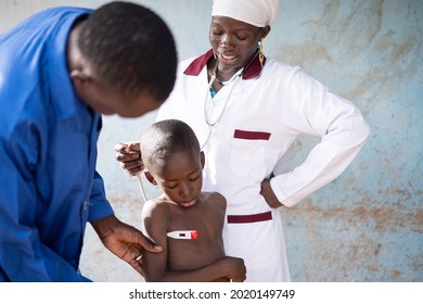 In This Image, A Young Black Nurse Is Talking To A Worried Tiny African Boy Undergoing Temperature Screening With A Digital Thermometer During A Routine Health Check In A Rural Hospital