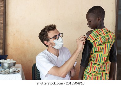 In This Image, A Young White Doctor Wearing A Protective Mask Is Disinfecting The Arm Of A Small Black Boy With A Cotton Ball Before Injecting Him A Vaccine During An Immunization Programme In Africa