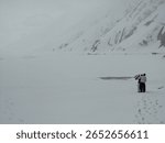 This impactful image portrays two figures standing on a vast, snow-covered frozen lake, set against the backdrop of snow-laden mountains under a cloudy sky in Pakistan.
