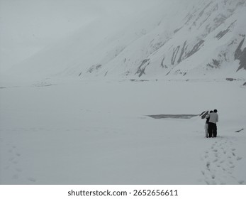 This impactful image portrays two figures standing on a vast, snow-covered frozen lake, set against the backdrop of snow-laden mountains under a cloudy sky in Pakistan.