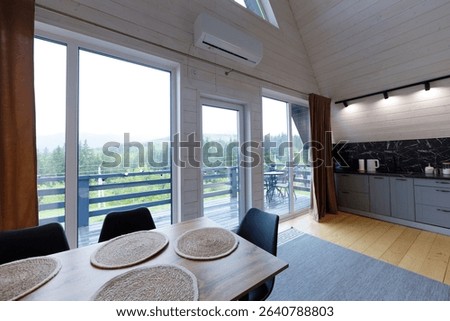 This inviting interior showcases a stylish dining area with natural touches, large windows bringing in scenic views of the green forest outside, and a functional kitchen space.