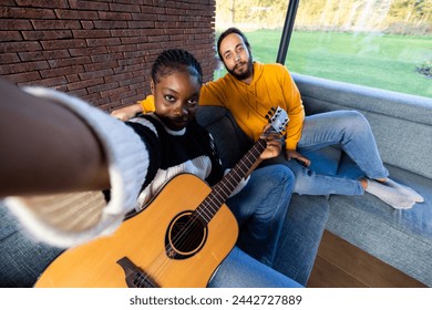 In this inviting scene, two friends share a moment of music and companionship. A woman, extending her arm for a selfie, holds a guitar, while a man beside her relaxes on the couch. The sunlit room and - Powered by Shutterstock