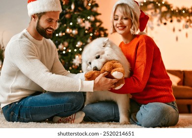 In this joyous holiday scene, a cheerful couple, donning festive Santa hats, joyfully smile while their fluffy dog happily joins them, all enveloped in a warm atmosphere of delightful decorations - Powered by Shutterstock