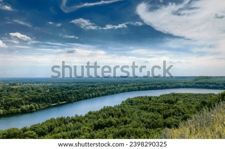 This landscape photograph offers a breathtaking panoramic view of a meandering river cutting through a lush green forest. The expansive blue sky, adorned with wispy clouds, crowns the scene