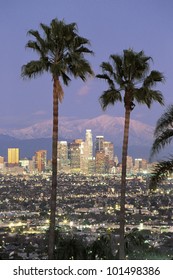 This is the Los Angeles skyline with two palm trees in the winter at dusk. Snowy Mount Baldy is in the background.