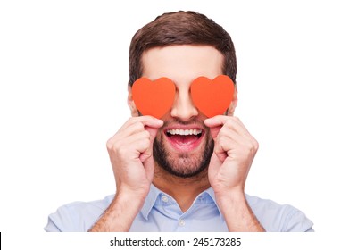 This is love! Handsome young man holding heart shaped valentine cards in front of his eyes and smiling while standing isolated on white background  