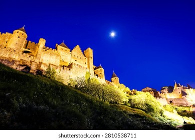 This low-angle shot captures the medieval fortress of Carcassonne, France, illuminated against a deep blue night sky with a bright moon overhead. The fortress walls and towers are bathed in warm light, contrasting with the cool tones of the sky and the dark, grassy hillside in the foreground.