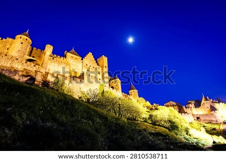 This low-angle shot captures the medieval fortress of Carcassonne, France, illuminated against a deep blue night sky with a bright moon overhead. The fortress walls and towers are bathed in warm light, contrasting with the cool tones of the sky and the dark, grassy hillside in the foreground.