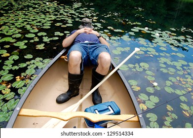  This Man Is Shown Dressed For A Fishing Expedition, With Fishing Pole, Tackle Box, Fishing Hat, And Rubber Boots, Asleep In The Back Of His Canoe.  Lily Pads Fill The Water Around The Boat.
