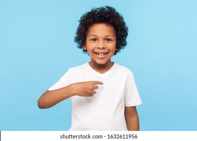 This is me. Portrait of happy preschool curly boy in T-shirt joyfully looking at camera and pointing to himself, proud of own success, egoistic child. indoor studio shot isolated on blue background