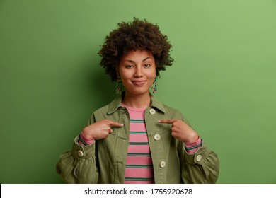 This is me! Satisfied proud Afro American woman points at herself, stands self confident, dressed fashionably, feels successful of her own achievement, stands indoor against green background