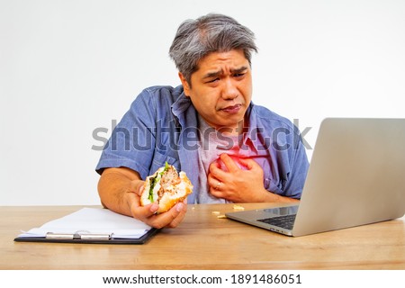 This middle-aged man eats a hamburger while sitting at work, laptop, computer, has chest pain, uncomfortable breathing, heart disease, touches his painful heart with his hands.
