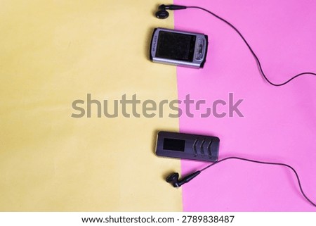 This minimalist composition features two vintage digital music players (MP3 players) mp4 from the early 2000s. The objects are placed on a two-tone surface (pink and yellow) with aesthetically pleasing earphone cables. It's suitable for tech nostalgia, the Y2K lifestyle, and retro gadget trends.