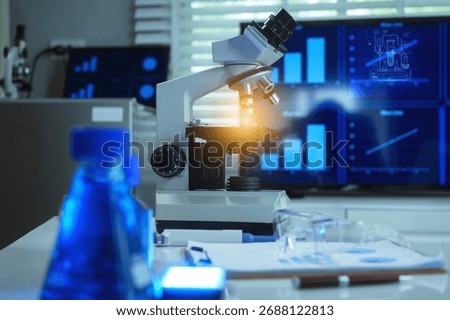 This modern laboratory setup features a brightly lit microscope on a desk, a blue chemical flask in the foreground, and dual screens displaying scientific research data charts.