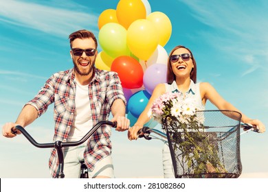 This is so much fun! Low angle view of cheerful young couple smiling and riding on bicycles with colorful balloons in the background