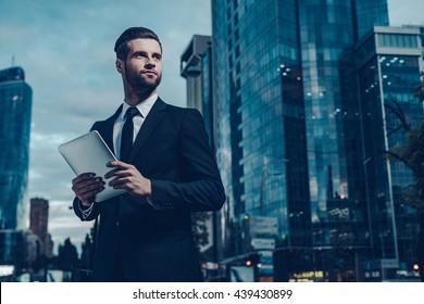 This Is My City. Night Time Image Of Confident Young Man In Full Suit Holding Digital Tablet And Looking Away While Standing Outdoors With Cityscape In The Background