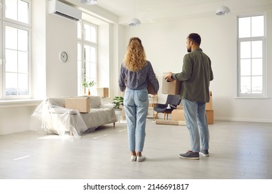 This is our home now. Young married couple settling down in new place. Rear view from behind of two people who are moving house or apartment standing in spacious room, holding boxes and looking around - Shutterstock ID 2146691817