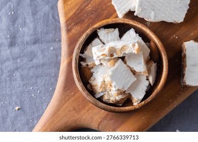This overhead shot features authentic Spanish turron de Alicante, a traditional holiday confection. The image displays broken pieces of the hard, white nougat packed with whole toasted almonds, presented in a round wooden bowl. The bowl sits atop a textured wooden serving board. Scattered around are larger rectangular bars of the same turron and another wooden dish containing raw almonds. The entire arrangement is set against a dark grey fabric background, highlighting the bright white color of the honey-based sweet and the warm wood tones.