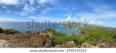 This panoramic image captures the famous view of English Harbour and Falmouth Harbour from the Shirley Heights lookout in Antigua.
The photo shows a high panoramic view of the blue Caribbean Sea and a winding coastline filled with green hills. Several white sailboats are anchored in the calm, turquoise waters of the harbor below. In the foreground, there are dark, jagged volcanic rocks and bright green tropical trees. The sky is a brilliant blue with soft, white clouds, showing a classic sunny day on the island.