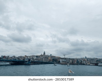 This panoramic shot captures the architectural splendor of Istanbul, highlighting the ancient Galata Tower and the busy Galata Bridge over the Golden Horn. The dramatic cloudy sky adds a unique atmosphere to the urban landscape, filled with historic buildings and active water traffic.