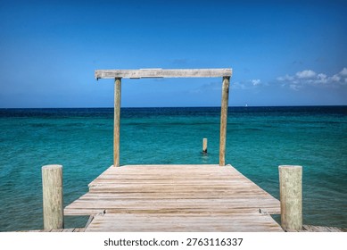 This peaceful scene features a rustic wooden pier stretching out over the vibrant turquoise Caribbean Sea, leading the eye toward the open ocean under a clear, brilliant blue sky.
A simple wooden frame structure stands at the end of the pier, creating a natural focal point that frames the expansive water and distant horizon.
Gentle ripples dance across the crystal-clear sea, which shifts in color from bright turquoise near the pier to deeper blue farther out.
A lone sailboat sails gracefully in the distance, enhancing the feeling of solitude, freedom, and timeless tropical serenity in this stunning coastal view.