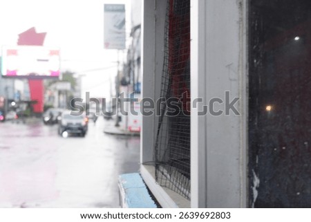 This photo captures a close-up of a worn security booth window with a bent metal mesh, set against a rainy urban street with blurred traffic and colorful billboards in the background during daylight.