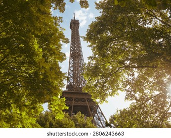 This photo captures the Eiffel Tower in Paris, framed beautifully by lush green trees. The tower rises majestically against a partly cloudy sky, with sunlight filtering through the leaves, creating a peaceful and inviting atmosphere. The scene highlights the iconic landmark amidst a vibrant, natural setting.