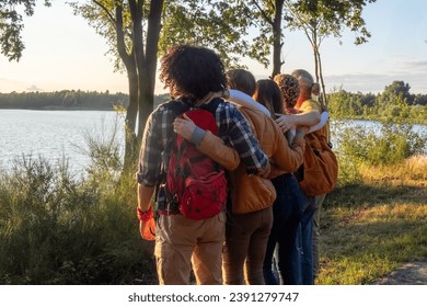 This photo captures a full-length rear view of five young, multiracial millennial friends as they celebrate reaching a summit near the shore of a forest lake. Bathed in the glow of the setting sun - Powered by Shutterstock