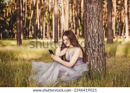 This photo depicts image of A beautiful young girl in a puffy gray dress sits by a tree on the grass in a park and uses her phone for correspondence, conversations, blogging, remote work and freelanc